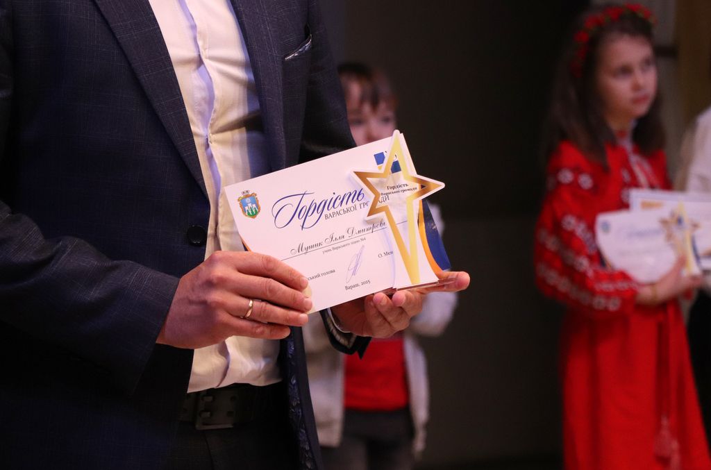 На передньому плані зображено чоловіка в темно-синьому костюмі, білій сорочці та краватці. Він тримає в руках грамоту та статуетку у формі зірки. На грамоті видно герб міста Вараш та напис "Гордість Вараської громади". Також видно підпис та дату "Вараш, 2025".   На задньому плані видно двох дітей. Дівчинка справа одягнена в червону вишиту сукню та вінок на голові, вона також тримає в руках подібну оформлену грамоту. Інша дитина стоїть трохи позаду та лівіше, її видно частково.