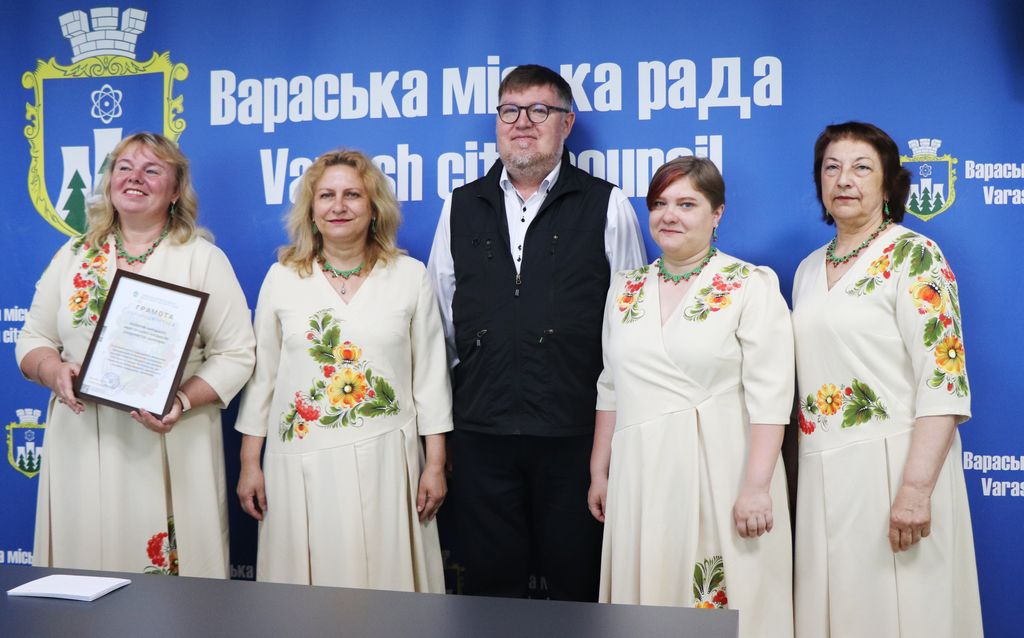 Олександр Мензул у чорному жилеті та окулярах стоїть по центру, оточений чотирма учасницями ансамблю у світлих сукнях з кольоровою вишивкою. Одна з жінок ліворуч тримає рамку з грамотою. Усі вони стоять на фоні синьої стіни з логотипами "Вараська міська рада" та "Varash city council".