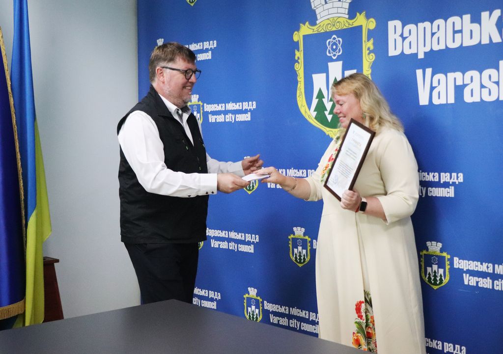 Олександр Мензул у чорному жилеті та білій сорочці вручає папери учаниці ансамблю у світлій сукні з вишитими елементами, яка тримає рамку з грамотою. На задньому плані — синя стіна з логотипами "Вараська міська рада" та "Varash city council".