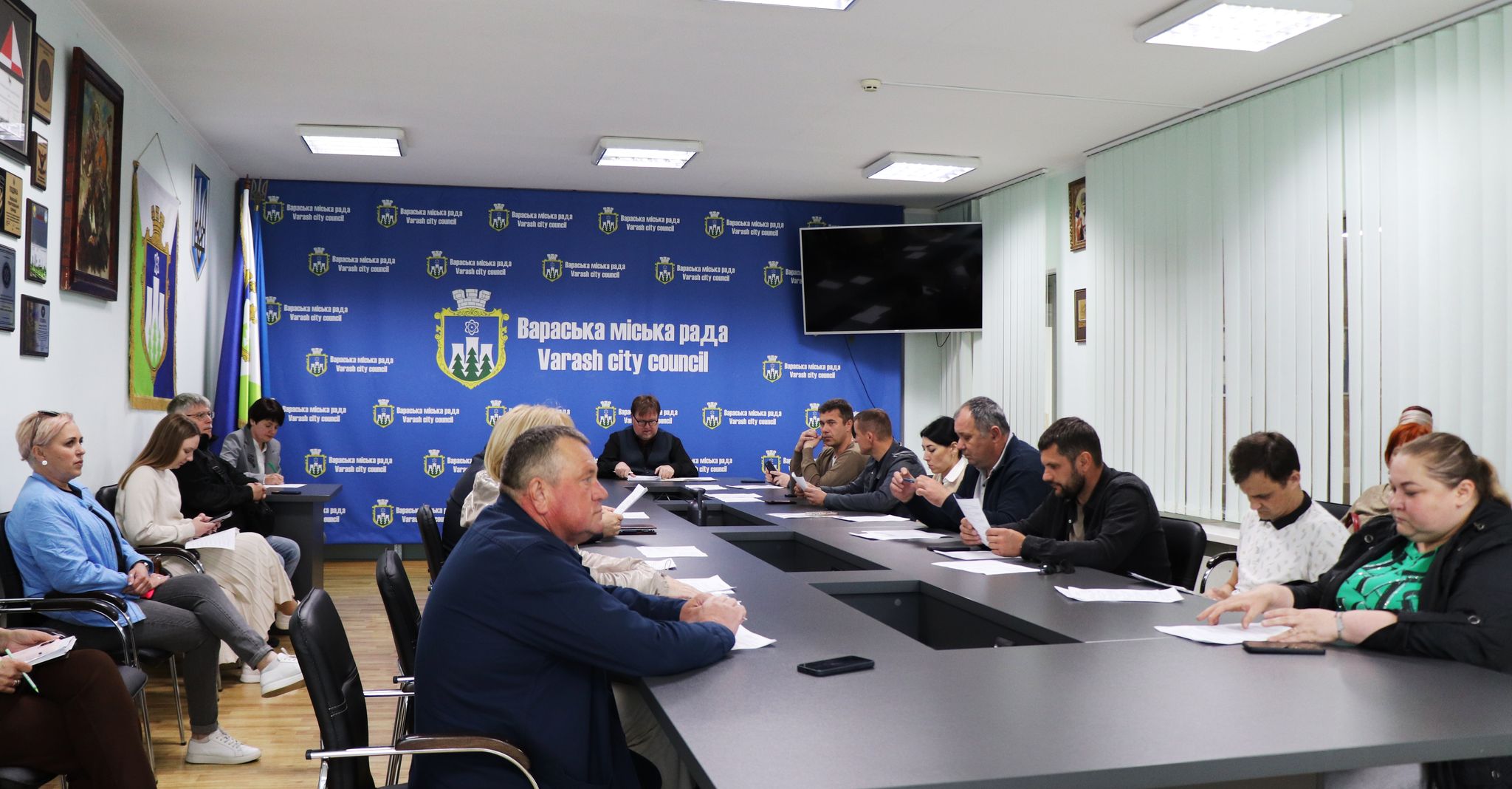 На фотографії зображено засідання виконавчого комітету Вараської міської ради. У просторій кімнаті за довгим столом, вкритим темно-сірою поверхнею, сидять люди. На чолі столу, ближче до центру кадру, сидить міський голова Вараша Олександр Мензул. Він уважно дивиться на когось за кадром.  По обидва боки від нього та вздовж столу розмістилися інші члени виконавчого комітету. Серед присутніх як чоловіки, так і жінки різного віку, одягнені в повсякденний або діловий одяг. Вони зосереджено слухають, роблять нотатки або переглядають документи, що лежать перед ними на столі.  На задньому плані видно стіну, оформлену синім банером з логотипом та написом "Вараська міська рада Varash city council", що повторюється. Також на стіні розміщено герб України, прапор Вараша та, можливо, інші офіційні символи. Праворуч на стіні висить плаский телевізор. Освітлення в кімнаті яскраве, штучне, від стельових світильників.