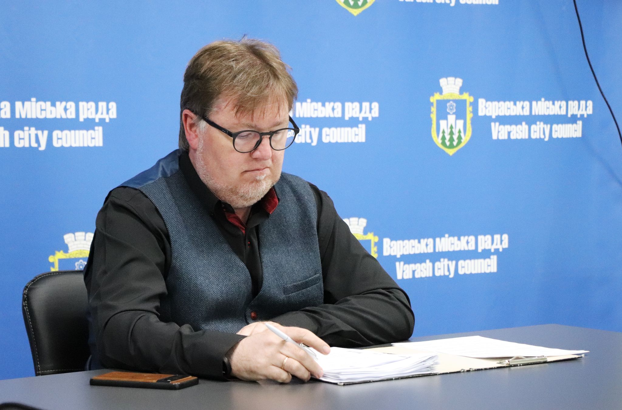 За столом сидить Олександр Мензул. Він середнього віку, має світле волосся з рудуватим відтінком, носить окуляри в темній оправі та темну жилетку поверх чорної сорочки з довгими рукавами. Він зосереджено дивиться на документи, що лежать перед ним на темному столі, і тримає в руці ручку. На столі також лежить мобільний телефон у коричневому чохлі. На задньому плані видно синій фон з повторюваним логотипом та написом "Вараська міська рада Varash city council".