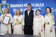 Олександр Мензул у чорному жилеті та окулярах стоїть по центру, оточений чотирма учасницями ансамблю у світлих сукнях з кольоровою вишивкою. Одна з жінок ліворуч тримає рамку з грамотою. Усі вони стоять на фоні синьої стіни з логотипами "Вараська міська рада" та "Varash city council".