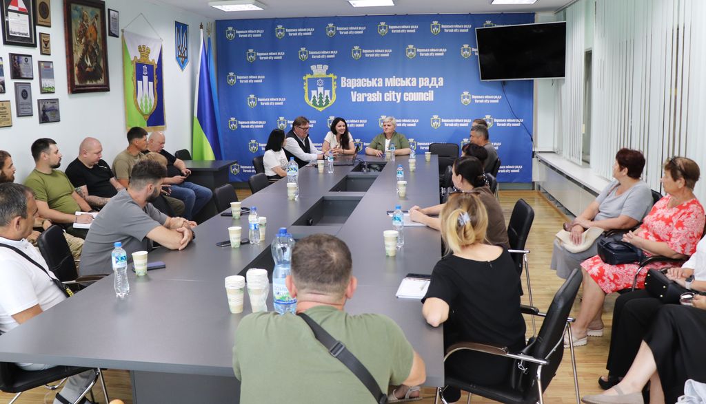 Широкий знімок залу, де група людей сидить за великим сірим столом під час зустрічі. На стіні позаду видно банер з написом "Вараська міська рада / Varash City Council".  На столі розставлені пляшки з водою та паперові стаканчики. На стінах також є прапори та інші елементи декору.