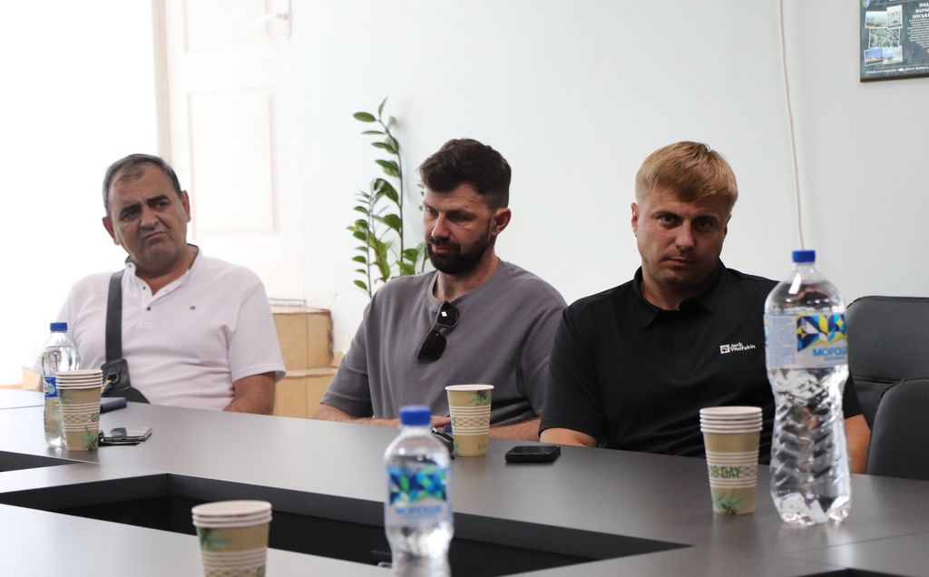 Троє чоловіків сидять за столом, дивлячись на щось поза кадром. Чоловік ліворуч, з сивим волоссям, одягнений у білу сорочку-поло. Чоловік посередині, з темним волоссям і бородою, одягнений у сіру футболку. Чоловік праворуч, зі світлим волоссям, одягнений у чорну сорочку-поло. На столі перед ними стоять пластикові пляшки з водою та паперові стаканчики.