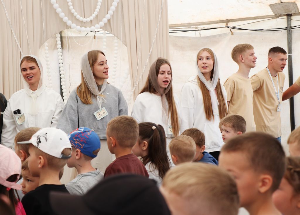 На фотографії зображена група молодих людей та дітей стоять у приміщенні: шатер. На передньому плані видно голови та спини дітей. На задньому плані стоїть група молодих людей. Деякі з дівчат мають на головах світлі хустки. Одна дівчина в білому одязі та хустці посміхається, інша, у сірій толстовці та світлій хустці, дивиться праворуч і також усміхається. Ще дві дівчини поруч також у світлому одязі та хустках. Поруч з ними стоять двоє молодих чоловіків, один з яких у бежевій футболці, а інший у світлій футболці з бейджем. На задньому плані видно світлі декорації, зокрема гірлянди з білих намистин.