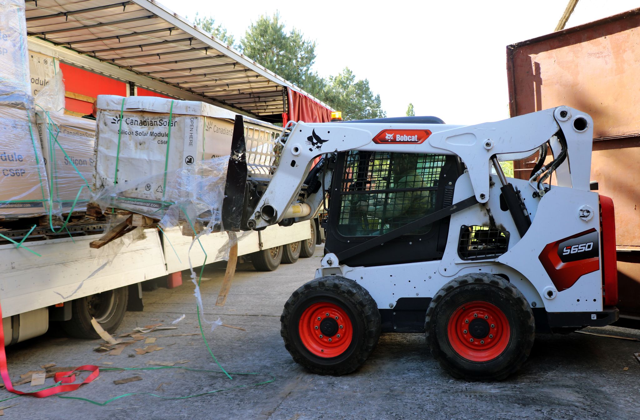 На зображенні показано білий колісний навантажувач Bobcat S650, який за допомогою вил підіймає велику картонну коробку з написом "Canadian Solar Silicon Solar Module CS6P" з кузова вантажівки.  Навантажувач стоїть на асфальтованій поверхні, частково в тіні від вантажівки. Вантажівка має білий кузов з тентом зверху, а її задня частина відкрита. У кузові видно інші подібні коробки з сонячними модулями.  На задньому плані видно зелені дерева та частково споруду з коричневими металевими воротами або стінами.  Навантажувач чітко маркований логотипом "Bobcat" та моделлю "S650". На коробці з сонячними модулями також є логотип "Canadian Solar".  Зображення фіксує момент розвантаження сонячних панелей з вантажівки за допомогою навантажувача Bobcat S650.