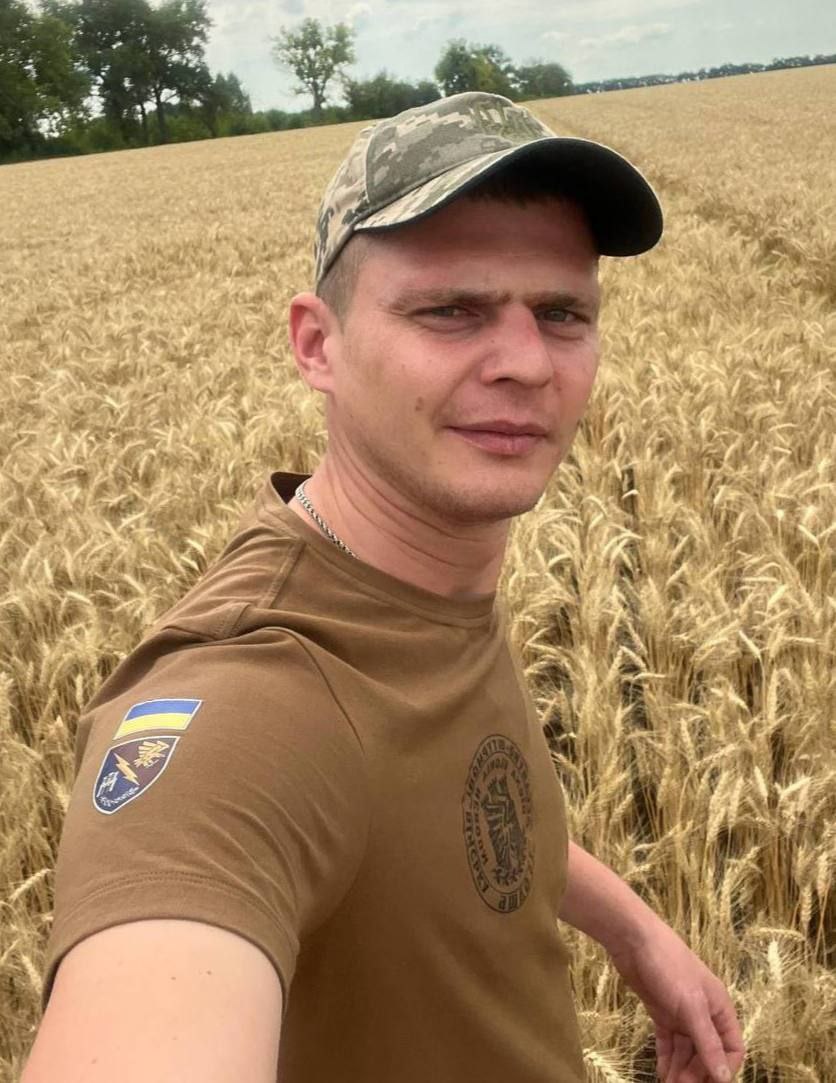 Портрет чоловіка, Віталія Гришка, у камуфляжній кепці та коричневій футболці з українським шевроном, який стоїть на пшеничному полі.