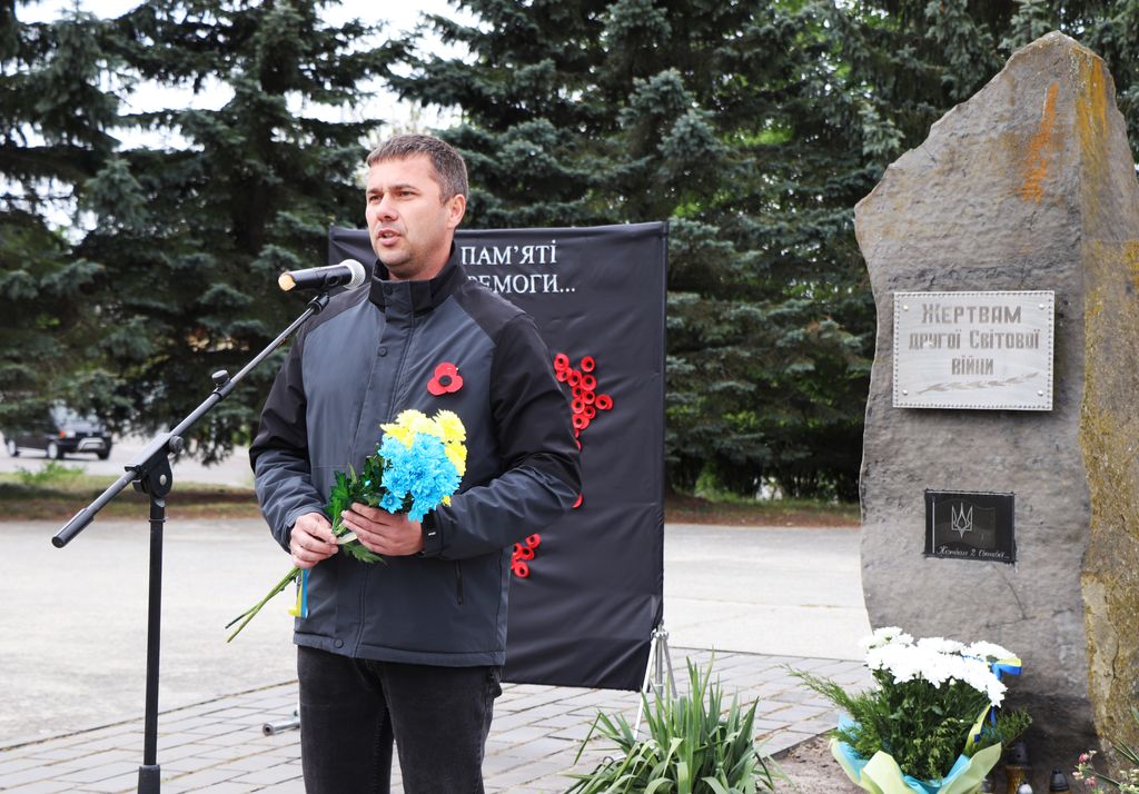 Заступник міського голови Роман Хондока стоїть біля мікрофона, тримаючи букет жовтих і блакитних квітів, на фоні меморіального знаку з написом "Жертвам Другої Світової Війни" та символікою України.