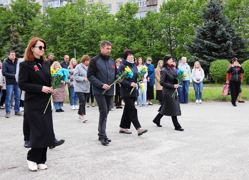 Група людей несе букети жовтих і блакитних квітів, йдучи тротуаром у напрямку пам’ятного знаку "Жертвам Другої світової війни". На одязі деяких людей видно червоні маки.