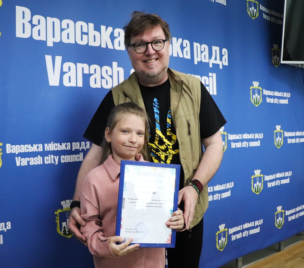 міський голова та нагороджена вихованка Вараської мистецької школи позують для фотографії, тримаючи документ у рамці, на тлі стіни з написом "Вараська міська рада".
