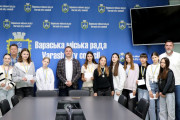 На фото зображена група людей у зеленій залі ВК ВМР. У центрі стоять юні дівчата, орієнтовно підліткового віку, які тримають у руках срібні медалі на синьо-жовтих стрічках.     По обидва боки від дівчат стоять двоє чоловіків старшого віку. Заступник міського голови Дмитро Стецюк стоїть ліворуч від центру, одягнений у сірий піджак у клітинку та світлу сорочку.     На задньому плані видно синю стіну з повторюваним логотипом та написом "Вараська міська рада" і "Varash City Council". Також на стіні розміщено герб Вараської міської ради. У правому верхньому кутку частково видно чорний екран.    Загальна атмосфера на фото урочиста та позитивна, що відповідає події нагородження.