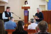 На зображенні видно сцену нагородження в приміщенні. У центрі, за дерев'яною трибуною з гербом України, стоїть жінка - начальник відділу персоналу виконавчого комітету Вараської міької ради, у світлій кофті та темних штанях, яка оголошує про нагородження. Праворуч від трибуни чоловік із сивим волоссям у темній сорочці та джинсах тримає в руках синю папку та зачитує з аркуша паперу. Ліворуч від трибуни стоїть міський голова міста Вараш Олександр Мензул, у світлій сорочці та темній жилетці, та аплодує. Перед сценою сидять слухачі. Задні ряди слухачів частково розмиті. За трибуною на стіні висить частина синьо-жовтого прапора.